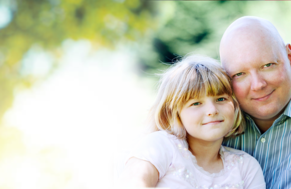 A bald man hugging his young daughter abd sitting outside in front of trees and the shining sunlight.